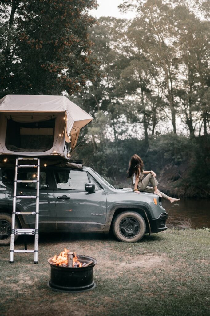 A woman sits on a jeep by a campfire, enjoying an outdoor adventure.